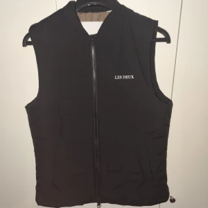 Svart väst från Les Deux - Säljer en stilren svart väst från Les Deux med dragkedja framtill. Västen har en enkel design med märkets logga broderad på bröstet. Perfekt för lager-på-lager under kyligare dagar. Står xs men passar bättre till m