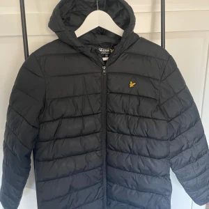 Svart dunjacka från Lyle & Scott - Säljer en svart dunjacka från Lyle & Scott med huva och dragkedja. Jackan har ett stilrent quiltat mönster och en liten gul logotyp på bröstet. Perfekt för kyligare dagar. Storleken är från 15-16 år eller runt S/M