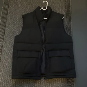 Svart dunväst från Saligia - Snygg svart dunväst från Saligia med dragkedja och stora fickor framtill. Perfekt för kyliga dagar när du vill hålla stilen. Västen har en klassisk design som funkar till det mesta. 🖤