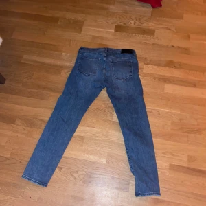 Blå jeans från Dressmann - Säljer ett par blå jeans från Dressmann i slim fit. De har en klassisk femficksdesign och slitna detaljer för en cool look. Perfekta för en avslappnad stil.
