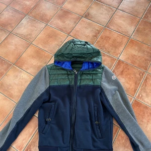 Moncler cardigan - Säljer en flerfärgad cardigan från Moncler med huva pga att den är för liten. Jackan har en kombination av grönt, blått och grått, är storlek M men sitter mer som S. Köptes i london för ca 6 år sedan men användes i en vår, sedan har den hängt i min garderob. Kom med förslag!