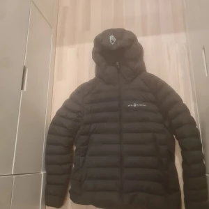 Svart dunjacka från Sailracing - Säljer en svart dunjacka från Sailracing med huva och dragkedja. Jackan har en quiltad design och är perfekt för lite kallare dagar men även under våren då den inte är för tjock. I mycket bra skick förutom ett hål som jag har lagat (bild 3) men inget som märks. 3200kr nypris mitt pris 500.
