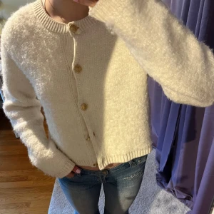 Vit stickad kofta med knappar - Mysig vit stickad kofta med träknappar framtill. Den har en rund halsringning och långa ärmar med ribbade muddar. 