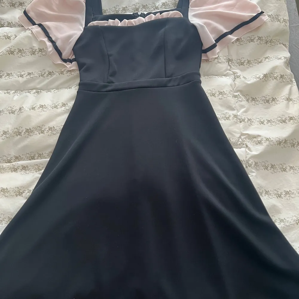 Elegant svart klänning med ljusrosa puffärmar och svarta detaljer. Klänningen har en figurnära passform upptill och en något utsvängd kjol. Perfekt för en stilfull look.. Mekot.