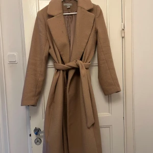 Beige kappa från H&M - Elegant beige kappa från H&M med knytbälte i midjan. Den har en klassisk krage och långa ärmar, perfekt för kyligare dagar. Stängs med bälte för en stilren look.