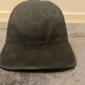 Svart keps från Gucci med GG-mönster - Snygg svart keps från Gucci med diskret GG-logga i hela tyget. Kepsen har böjd skärm och är tillverkad i ett slitstarkt material med klassisk passform. Perfekt accessoar för en trendig look.