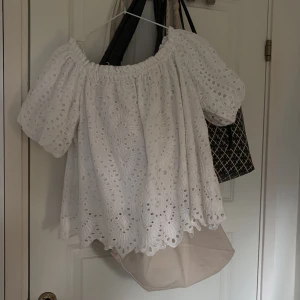 Vit blus med broderade detaljer - Superfin vit blus med broderade hålmönster och puffiga korta ärmar. Blusen har en offshoulder-modell med resår upptill och volangkant nertill. Perfekt för en somrig look!