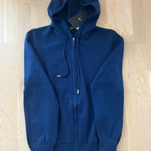 Mörkblå hoodie med dragkedja - Säljer en mörkblå hoodie med dragkedja och huva. Tröjan har långa ärmar, ribbade muddar och fickor framtill. Perfekt för en avslappnad stil och enkel att matcha med andra plagg.