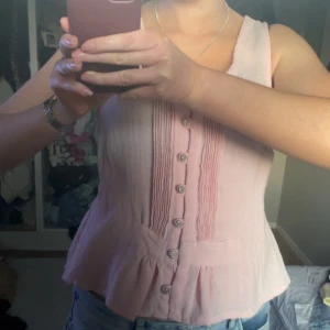 Rosa ärmlös blus med knappar - Säljer en söt rosa ärmlös blus med knappar framtill. Blusen har en v-ringad design och fina veckdetaljer på bröstet. Perfekt för varma dagar!