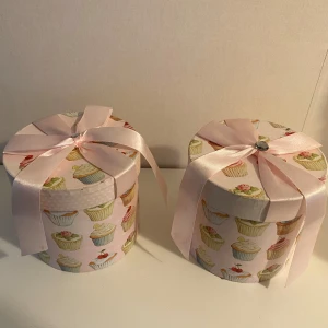 2 st Runda presentaskar med cupcakes-motiv och rosett - Säljer 2 söt rund presentask med pastellfärgat cupcakes-motiv och stor ljusrosa rosett på locket. Asken är ca 13 cm hög och 13 cm i diameter, perfekt för små presenter eller förvaring av smycken och småsaker.