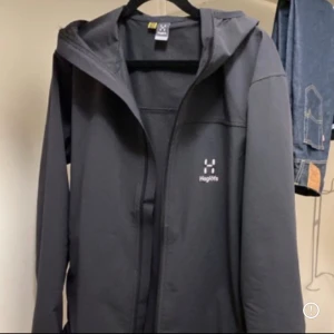 Svart softshelljacka med huva från Haglöfs - Svart softshelljacka från Haglöfs med huva och dragkedja framtill. Jackan har en stilren design med diskret logga på bröstet. Perfekt för utomhusaktiviteter och vardagsbruk.