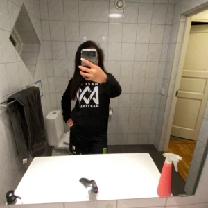 Svart hoodie från Marcus & Martinus - Svart hoodie med stor vit Marcus & Martinus-logga på bröstet. Tröjan har huva med dragsko och en stor magficka framtill. Perfekt för dig som gillar streetwear och vill ha något bekvämt och snyggt.