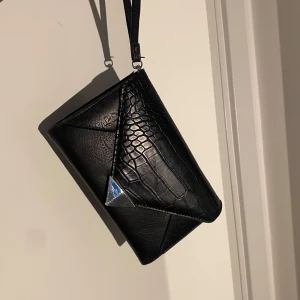 Svart axelväska med kedja och krokopräglat mönster - Axelremsväska köpt i fakebutik i Grekland. Bandet går att justera. Storlek 19 x 12 cm. Se bilder för defekter. 