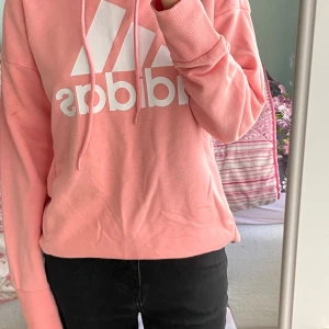 Rosa hoodie från Adidas - Säljer en snygg rosa hoodie från Adidas med stor vit logga framtill. Tröjan har huva med dragsko och ribbade muddar vid ärmslut och nederkant. Perfekt för en avslappnad och sportig stil.