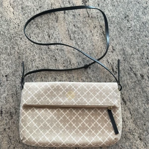 Beige axelväska från By Malene Birger - Snygg beige axelväska med vitt grafiskt mönster från By Malene Birger. Väskan har guldfärgad dragkedja, svart axelrem och en praktisk innerficka. Perfekt för att bära dina viktigaste saker med stil.