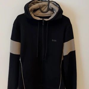 Svart hoodie med beige detaljer från BOSS - Säljer en svart hoodie från BOSS med beige paneler på ärmar och huva. Tröjan har dragkedja framtill, justerbar huva och diskret BOSS-logga på bröstet. Perfekt för en avslappnad och stilren look.