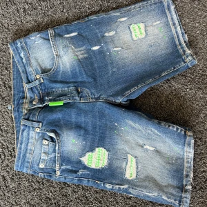 Blå slitna jeansshorts från MY BRAND - Säljer ett par blå jeansshorts från MY BRAND med slitna detaljer, gröna inslag och patchar. Shortsen har normal passform, klassisk femficksdesign och gröna detaljer på fickorna. Perfekta för en avslappnad stil.