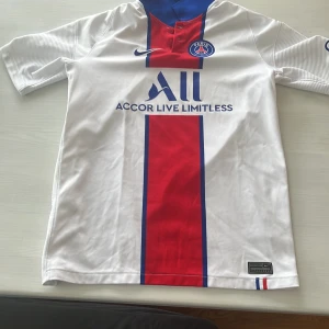 PSG hemmatröja 2020/21 - Vit PSG fotbollströja med röd och blå vertikal rand, blå Nike-logga och klubbmärke på bröstet. Kragen är blå och tröjan har sponsortryck på framsidan. Klassisk design från säsongen 2020/21.