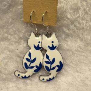 Vita och blå kattörhängen - Säljer ett par unika örhängen formade som katter i vit och blå färg. Örhängena har ett fint bladmönster och är perfekta för dig som gillar djur och lekfull design. Hängande modell med krok.