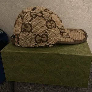 Beige keps från Gucci med GG-mönster - Snygg beige keps från Gucci med klassiskt brunt GG-mönster över hela kepsen. Justerbar rem baktill i brunt läder och böjd skärm. Perfekt accessoar för en trendig look.