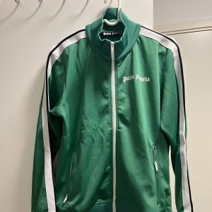 Grön track jacket från Palm Angels - Säljer en grön track jacket från Palm Angels med vit dragkedja och vita ränder längs ärmarna. Jackan har hög krage, två fickor med dragkedja och logga tryckt på bröstet. Perfekt för en sportig och trendig look. Helt oanvänd!