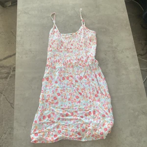 Blommig sommarklänning med smala axelband - Säljer en söt blommig sommarklänning med smala axelband. Klänningen har ett färgglatt mönster i rosa, blått och gult på en vit bakgrund. Den är smockad upptill och har en luftig kjol som ger en avslappnad känsla. Perfekt för varma dagar!