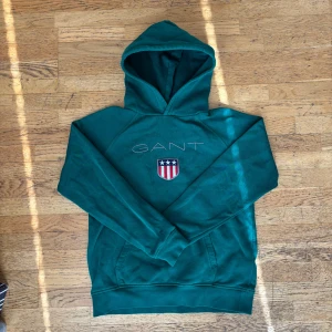 Grön hoodie från GANT - Säljer en snygg grön hoodie från GANT med broderad logga och emblem på bröstet. Tröjan har en klassisk känguruficka och en bekväm huva. Perfekt för en avslappnad stil.