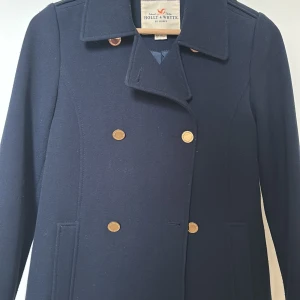 Mörkblå kappa från Holly & Whyte - Elegant mörkblå kappa från Holly & Whyte by Lindex med dubbelknäppning och stilrena guldknappar. Perfekt för en klassisk look. Kappan har långa ärmar och en tidlös design som passar till många tillfällen.