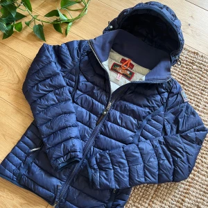 Mörkblå dunjacka från Parajumpers - Säljer en mörkblå dunjacka från Parajumpers med huva och dragkedja. Jackan är superlätt och har ett broderat märke på ärmen. Perfekt för kyliga dagar och har en sportig look med snygga detaljer.
