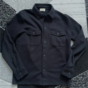 Svart overshirt från Selected Homme - Stilren svart overshirt från Selected Homme i ekologiskt material. Skjortan har två bröstfickor med lock och knappar framtill. Perfekt att bära som lager över en t-shirt eller tröja. Sitter bra, pris kan diskuteras.