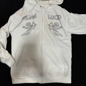 Vit hoodie med strasstenar från Bling Lucid - Vit hoodie med dragkedja från Bling Lucid. På bröstet finns texten 'BLING' och 'LUCID' i strasstenar samt två änglar i samma stil. Jackan har huva och fickor framtill. Perfekt för dig som gillar unika detaljer och streetwear. Pris kan diskuteras