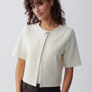 Vit kortärmad kofta med knapp - Stilren vit kofta med korta ärmar och rund halsringning. Jackan har en elegant guldig knapp framtill. Perfekt för en dressad look.