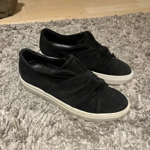 Svarta sneakers med knytdetalj från Rizzo - Säljer ett par svarta sneakers från Rizzo med snygg knytdetalj framtill. Skorna har vit platt sula och är tillverkade i mocka. Sparsamt använda. 