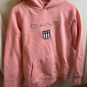 Rosa hoodie från GANT - Säljer en rosa hoodie från GANT med broderad logga och emblem på bröstet. Tröjan har huva och en stor magficka framtill. Perfekt för en avslappnad stil.