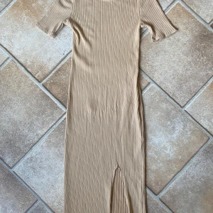 Beige ribbad figursydd klänning Gina Tricot  - Snygg beige ribbad klänning från GT med korta ärmar och en stilren slits framtill. Perfekt för en avslappnad men chic look. Klänningen har en kroppsnära passform och är gjord i ett mjukt material. Använd 2-3 gånger. Topp skick. 