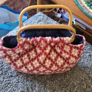 Rutig handväska med bambuhandtag - Charmig handväska med rött och vitt ulltyg rutmönster och bambuhandtag. Väskan har en mjuk, vadderad design och en mörkblå insida. Perfekt för en unik och stilfull look.