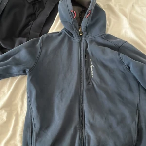 Blå softshelljacka från Sail Racing - Snygg blå softshelljacka från Sail Racing med dragkedja framtill och en praktisk bröstficka. Jackan har en hög krage och långa ärmar, perfekt för kyligare dagar. Den är stilren och funktionell, idealisk för både vardag och utomhusaktiviteter.