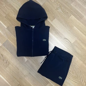 Lacoste Tracksuit - Mörkblå - Säljer en stilren mörkblå hoodie från Lacoste med dragkedja och huva. Tröjan har långa ärmar och en liten Lacoste-logga på bröstet. Perfekt för en avslappnad look.                                                Koftan saknar snöre i luvan och är i god skick, använd under en period men ren och fin fortfarande.                                                                      Storlek: S