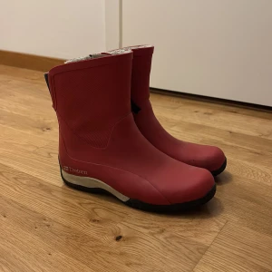 Röda gummistövlar från Tretorn - Säljer ett par röda gummistövlar från Tretorn med svart sula och vit insida. Stövlarna har dragkedja på sidan och är perfekta för regniga dagar. Klassisk design med rund tå och slitstark sula.