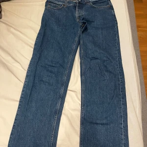 jeans från Nudie Jeans i modellen Rad Rufus strl:34/34 - Säljer ett par klassiska blå jeans från Nudie Jeans med raka ben och snygga kontrastsömmar. Byxorna har fem fickor, gylf med dragkedja och Nudie-logga på bakfickan. Perfekta för dig som gillar en tidlös jeansstil.
