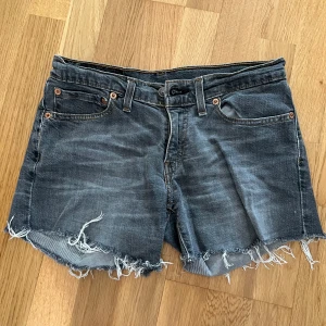 Grå jeansshorts med fransar från Levi's - Säljer ett par grå jeansshorts från Levi's med råa fransade kanter och klassisk femficksdesign. Shortsen har bälteshällor och knappgylf. Perfekta för en avslappnad stil.
