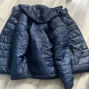 Svart quiltad dunjacka från Jack & Jones - Säljer en svart quiltad dunjacka från Jack & Jones med huva och rödfärgat innerfoder. Jackan har dragkedja och två sidofickor. Perfekt för kyliga dagar och enkel att matcha med olika outfits.