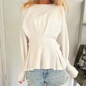 Vit blus med veckade detaljer - Säljer en stilren vit blus med unika veckade detaljer framtill och längs ärmarna. Blusen har lång ärm och en lätt utsvängd midja som ger en snygg siluett. Perfekt att matcha med jeans eller kjol.