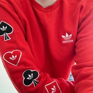 Röd långärmad sweatshirt från Adidas - Röd sweatshirt från Adidas med vita broderade loggor på bröstet och spelkortsmärken längs ärmarna. Tröjan har rund halsringning och är tillverkad i mjukt bomullsmaterial. Perfekt för en sportig och avslappnad stil. Orginalpris 800kr. Använd 1 gång. S och kan bäras i XS
