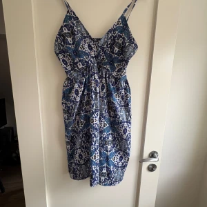 Blå- och vitmönstrad sommarklänning med smala axelband - Säljer en blå- och vitmönstrad klänning med smala axelband och lätt rynkad midja. Klänningen har ett bohemiskt mönster och är perfekt för varma dagar. Den är ärmlös och har en luftig passform som gör den bekväm att bära. Aldrig använd. 