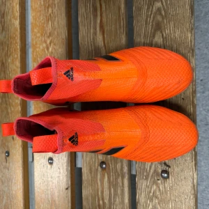 Orange fotbollsskor från Adidas - Säljer ett par riktigt snygga orangea fotbollsskor från Adidas med svarta ränder på sidan. Skorna har en strumpliknande passform och fasta dobbar för spel på gräs. Perfekta för dig som vill sticka ut på planen!   De är ett par Adidas ace 17+ samma som özil och pogba spelade med.