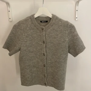 Grå stickad kofta från Gina Tricot - Kortärmad, grå stickad kofta från Gina Tricot med rund halsringning och knappar framtill. Perfekt för lager-på-lager och enkel att matcha med olika outfits. Köpte för 400 kr och nästan aldrig andvänd 
