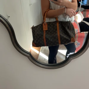 Louis Vuitton Keepall Monogram väska - Säljer en klassisk Louis Vuitton Keepall väska i brunt monogrammönster med detaljer i ljusbrunt skinn. Väskan har två handtag, dragkedja och ikoniska LV-loggor över hela ytan. Perfekt storlek för weekendresor och har en tidlös design. Den är köpt för flera år sedan i Paris i LV butiken. Men tyvärr pga av åldern finns inget äkthetsbevis. Den är också lite sliten pga åldern men utifrån är den overall supersnygg och fin. 