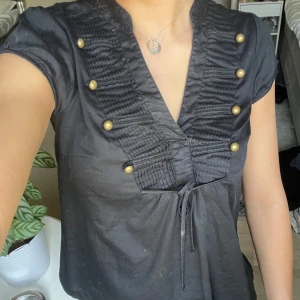 Svart blus med guldknappar och knyt - Svart blus med korta ärmar, dekorativa guldknappar framtill och plisserade detaljer vid bysten. Djup v-ringning och knytband i midjan. Perfekt för en trendig och stilren look.
