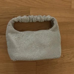Glittrig silver handväska - Säljer en snygg glittrig handväska i silver med ett unikt handtag som har rynkad design. Perfekt för att lyfta din outfit med lite extra bling. Väskan har dragkedja upptill och är kompakt i modellen.